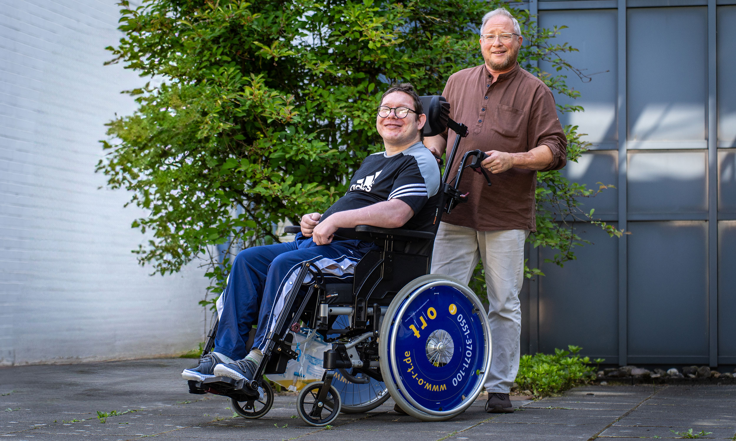 Pflegekraft zusammen im Portr�t mit einem jungen Pflegebed�rftigen im Rollstuhl vor der Einrichtung